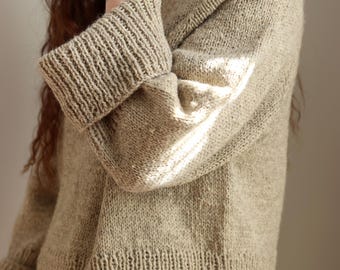 May include: A beige knit sweater featuring a high ribbed neckline, long sleeves with wide ribbed cuffs, and a ribbed hemline with side slits. The garment is crafted from a textured, heathered yarn in a neutral light brown tone with a visible knit pattern.