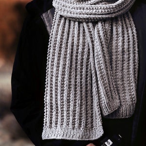 A man wears a ribbed knitted scarf, The Mountaineer Scarf. This gray knit mens scarf is a fashion scarf great for fall and winter. [knitted scarves, men knit scarf, rib scarf knitting pattern, beginner scarf knit patterns, beginner knit pattern]