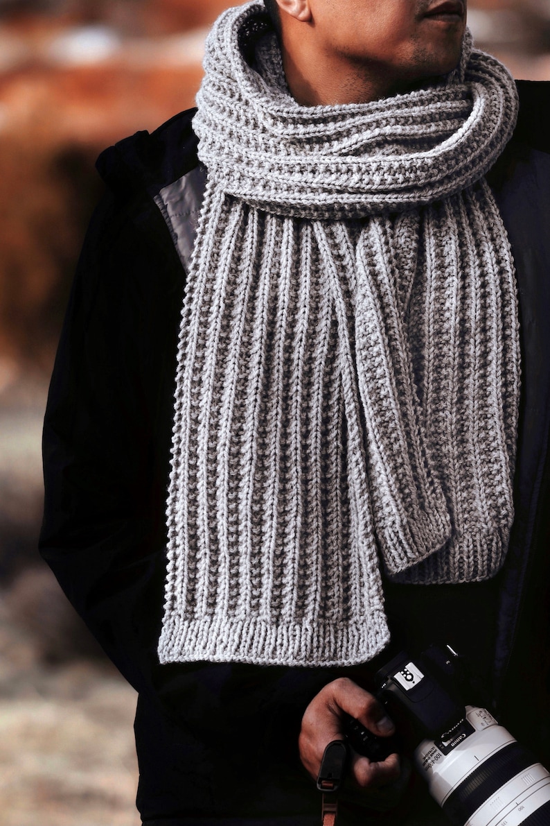 A man wears a ribbed knitted scarf, The Mountaineer Scarf. This gray knit mens scarf is a fashion scarf great for fall and winter. [knitted scarves, men knit scarf, rib scarf knitting pattern, beginner scarf knit patterns, beginner knit pattern]