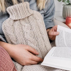 May include: A beige hot water bottle cover featuring a vertical cable knit pattern and a ribbed turtleneck collar. The cover is crafted from a thick, textured yarn and is designed to fit snugly around a standard rectangular hot water bottle.