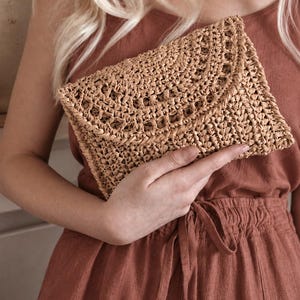 Può includere: Una pochette rettangolare realizzata in rafia intrecciata color marrone chiaro. La borsa presenta una trama testurizzata a maglia aperta con una patta che si ripiega sul davanti. Le fibre naturali creano una tonalità terrosa e uniforme.