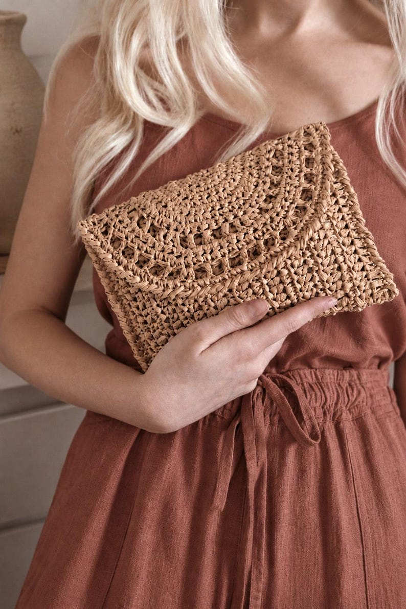 May include: A rectangular clutch bag crafted from woven tan straw material. The accessory features a detailed, intricate openwork pattern across the front flap and body, creating a textured, natural appearance with a consistent light brown color.