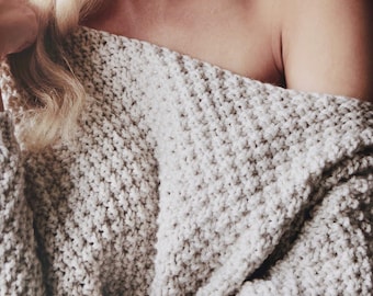 May include: A beige, chunky knit sweater featuring a wide, off-the-shoulder neckline. The garment is crafted with a thick, textured yarn in a consistent, repeating stitch pattern that creates a dense, cozy appearance across the entire fabric.