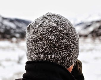 May include: A knit beanie featuring a heathered gray and white speckled pattern with a ribbed texture. The hat is worn over a black high-neck jacket. The knit construction shows a consistent weave throughout the crown and the folded brim edge.