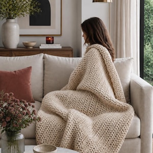 May include: A chunky, cream-colored knit throw blanket with a visible, textured stitch pattern. The blanket is draped over a light-colored sofa, covering the seat and back cushions, featuring a thick, soft yarn construction throughout.