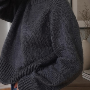 Pode incluir: Uma camisola de manga comprida em cinzento-carv&atilde;o escuro com gola redonda grossa e canelada, punhos canelados e uma bainha larga canelada. A pe&ccedil;a de malha tem um corte descontra&iacute;do com ombros desca&iacute;dos e uma textura de fio grosso.
