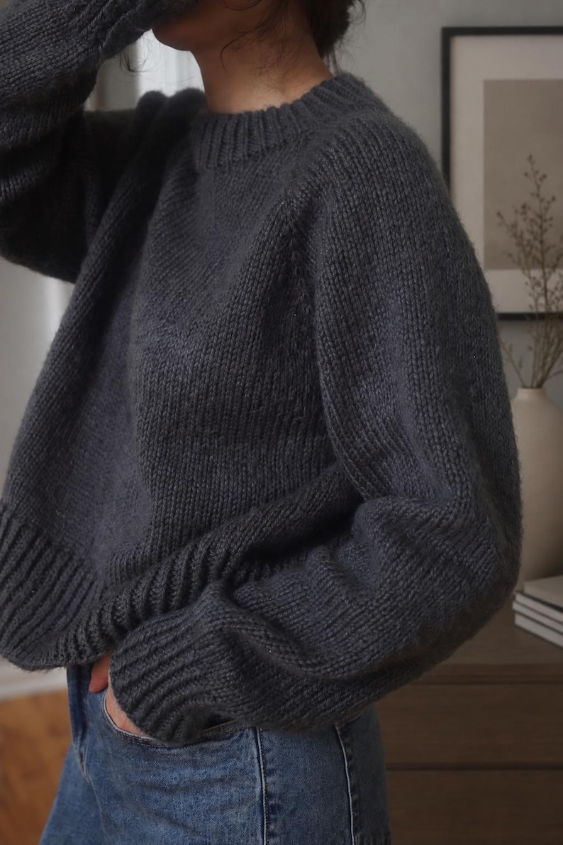 Pode incluir: Uma camisola de manga comprida em cinzento-carv&atilde;o escuro com gola redonda grossa e canelada, punhos canelados e uma bainha larga canelada. A pe&ccedil;a de malha tem um corte descontra&iacute;do com ombros desca&iacute;dos e uma textura de fio grosso.