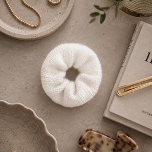May include: A white knitted fabric hair scrunchie centered among various accessories including gold-toned rings, a gold chain necklace, a tortoiseshell patterned hair claw clip, two gold hair pins, and a book with visible text on the cover.