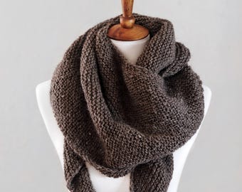 May include: A dark brown triangular scarf featuring a chunky garter stitch knit texture. The scarf is draped in multiple layers around the neck, showcasing a thick, consistent yarn construction with a visible weave pattern throughout the entire piece.
