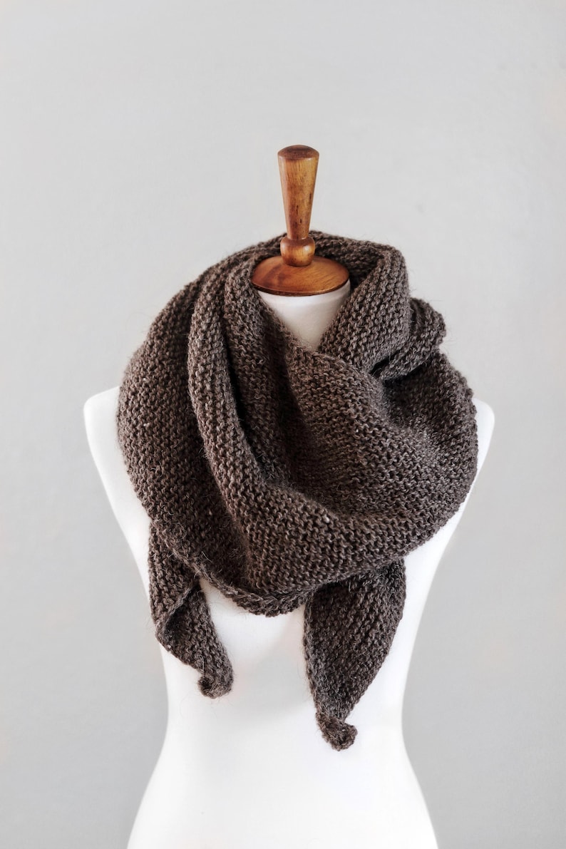 May include: A dark brown triangular scarf featuring a chunky garter stitch knit texture. The scarf is draped in multiple layers around the neck, showcasing a thick, consistent yarn construction with a visible weave pattern throughout the entire piece.