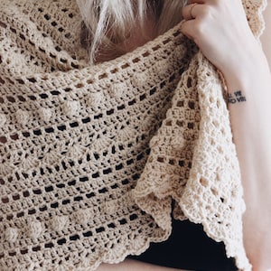 Women wears a crochet shawl scarf, The Isla Scarf, that features lace and bobbles. This triangle shawl scarf was crocheted in a cream yarn. [crochet shawl patterns, crochet scarf pattern, bobble crochet scarf, triangle crochet scarf pattern]