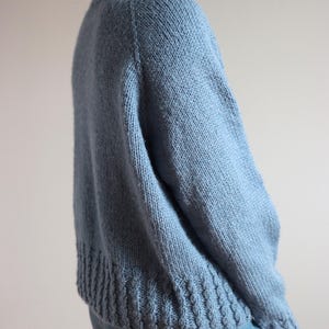 Cable knit sweater pattern, easy raglan sweater knitting pattern. women top down seamless raglan sweater with cables. oversize knit sweater pattern for women. beginner turtleneck pullover sweater knit pattern. high neck jumper knit pattern mock neck. A light blue knit sweater with a ribbed hem and cuffs. The sweater has a crew neck and long sleeves. The knitwear is a solid color and appears to be made of a soft, warm material. The sweater is paired with blue jeans.