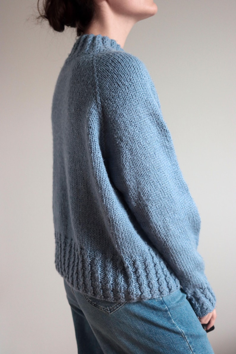 Cable knit sweater pattern, easy raglan sweater knitting pattern. women top down seamless raglan sweater with cables. oversize knit sweater pattern for women. beginner turtleneck pullover sweater knit pattern. high neck jumper knit pattern mock neck. A light blue knit sweater with a ribbed hem and cuffs. The sweater has a crew neck and long sleeves. The knitwear is a solid color and appears to be made of a soft, warm material. The sweater is paired with blue jeans.