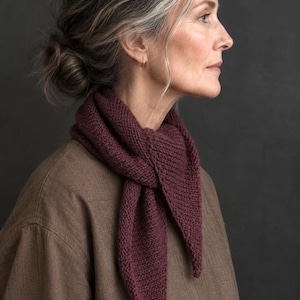 May include: A deep burgundy triangular knit scarf featuring a textured garter stitch pattern. The scarf is tied in a simple knot at the front, with two pointed ends hanging down over a plain brown long-sleeved shirt made of a woven fabric.