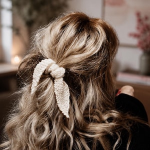 May include: A cream-colored knitted hair accessory featuring a knotted design with two dangling fabric tails. The accessory is secured around a section of wavy, highlighted hair, creating a textured look with its visible yarn loops and soft finish.