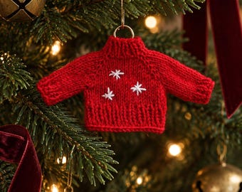 May include: A miniature red knitted sweater ornament featuring a ribbed mock neck, long sleeves, and a ribbed hem. The front of the sweater is decorated with three small, white embroidered star-shaped patterns. A gold metal ring is attached to the collar.
