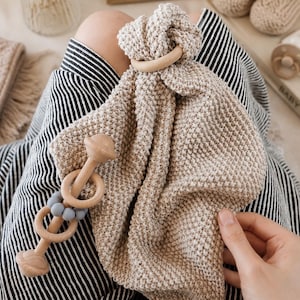 May include: A beige knitted security blanket featuring a smooth wooden ring at the top, paired with a wooden rattle toy that includes two wooden rings and three small grey silicone beads on a central wooden rod with rounded ends.