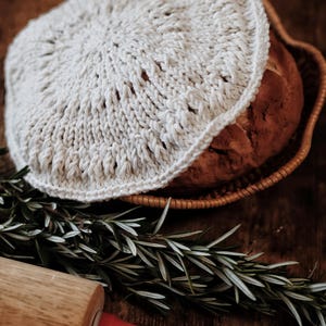 May include: A loaf of bread in a woven basket, covered with a cream-colored, crocheted bread cover. A wooden rolling pin with a red handle and sprigs of rosemary are on a dark wooden surface.