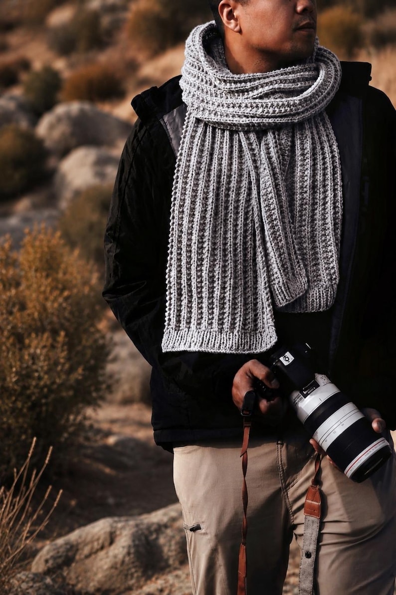 May include: A long, light gray knitted scarf with a vertical ribbed texture, worn wrapped around the neck. The scarf features a consistent, repeating stitch pattern throughout its length, ending in a simple, straight-edged hem at the bottom.