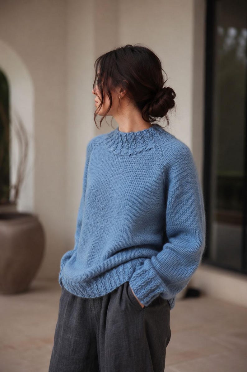 Cable knit sweater pattern, easy raglan sweater knitting pattern. women top down seamless raglan sweater with cables. oversize knit sweater pattern for women. beginner turtleneck pullover sweater knit pattern. high neck jumper knit pattern mock neck.  A light blue knitted sweater with a high neckline and textured details. The sleeves have a thumbhole design and the cuffs and hem feature a cable knit pattern. The sweater is made of a soft, possibly wool, material.