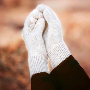 May include: A pair of white knit gloves featuring a soft, fuzzy texture and a ribbed cuff design. The gloves are constructed with a classic five-finger silhouette, showcasing a clean, solid white color throughout the entire knitted fabric.