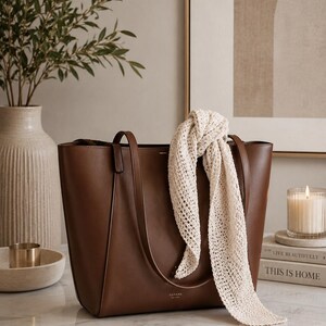 May include: A structured brown leather tote bag with two shoulder straps and the brand name CUYANA NEW YORK printed in gold on the front. A long, cream-colored, open-knit scarf is draped over one of the bag's handles, hanging down the front.