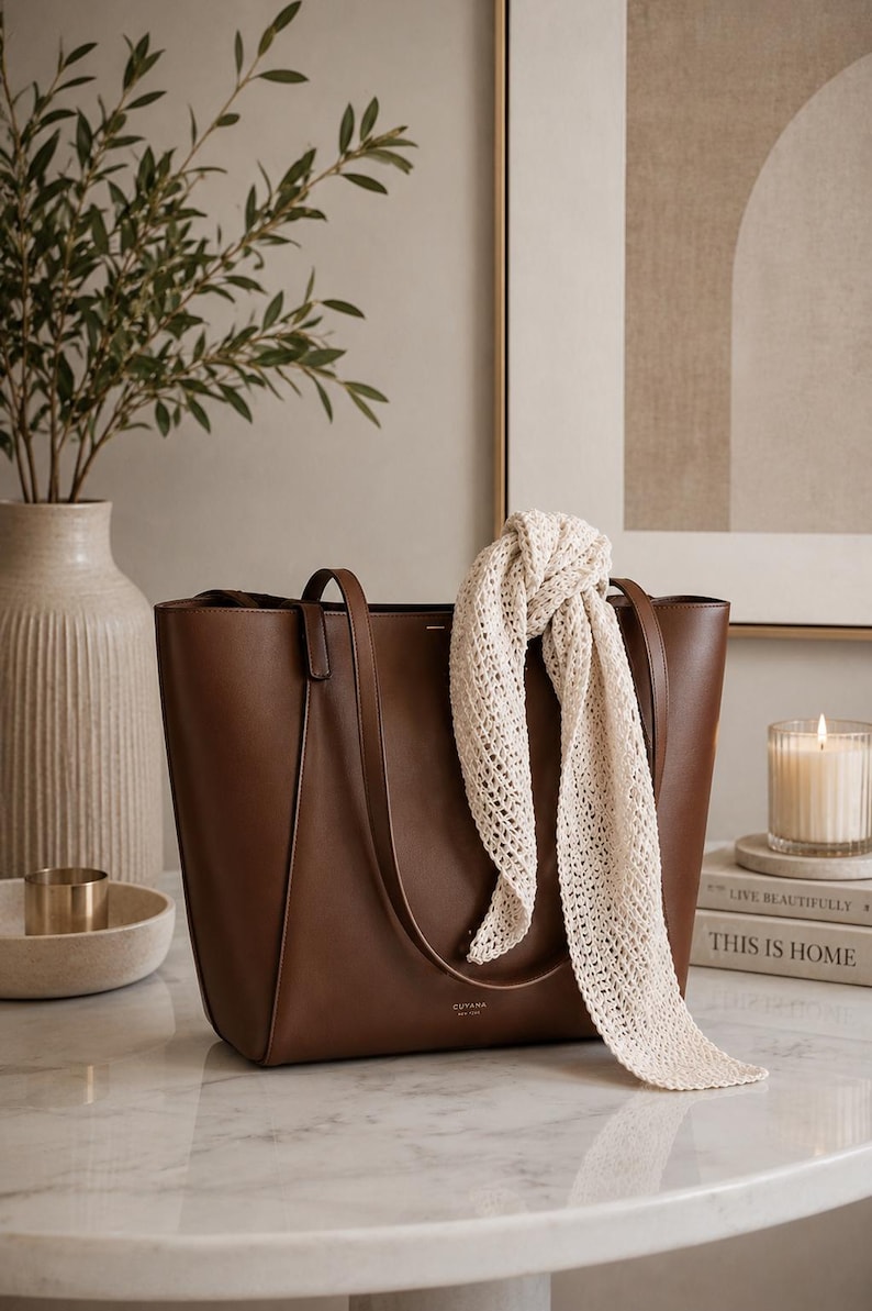 May include: A structured brown leather tote bag with two shoulder straps and the brand name CUYANA NEW YORK printed in gold on the front. A long, cream-colored, open-knit scarf is draped over one of the bag's handles, hanging down the front.