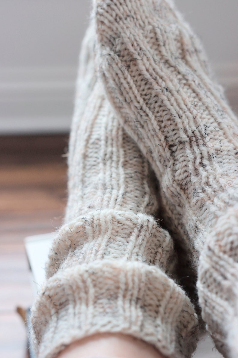 May include: A pair of thick, hand-knitted socks in a light beige, oatmeal-colored yarn. The socks feature a prominent vertical ribbed texture throughout the fabric, with subtle dark flecks of fiber woven into the light-colored wool blend material.