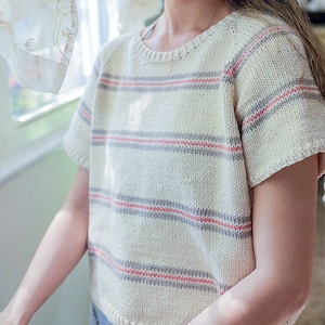 Woman wears a basic knit tee shirt, The Classic Tee, in cream with stripes. This simple knitted tshirt looks lightweight and great as a summer top. This easy tshirt features ribbing and classic knit stitches.
[sweater knitting patterns, easy knit]