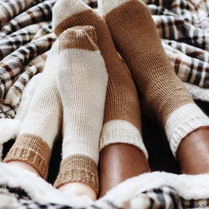 Könnte beinhalten: Zwei Paare brauner und weißer gestreifter Socken werden von zwei Personen getragen, deren Füße unter einer karierten Decke versteckt sind.