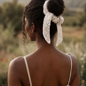 May include: A cream-colored crochet hair accessory tied around a bun, featuring an openwork stitch pattern with two long, tapered ends that hang down. The accessory is made of a textured yarn and has a knotted center detail securing it to the hair.
