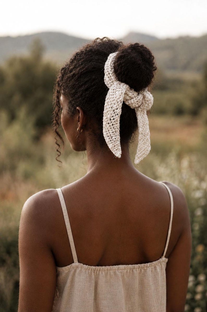 May include: A cream-colored crochet hair accessory tied around a bun, featuring an openwork stitch pattern with two long, tapered ends that hang down. The accessory is made of a textured yarn and has a knotted center detail securing it to the hair.