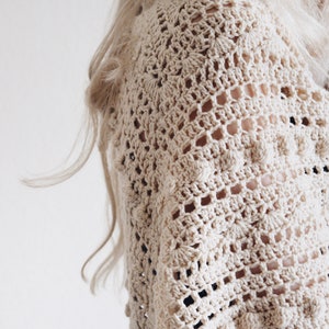 Woman wears a lace and bobble crochet scarf, The Isla Scarf, which was crocheted in cream yarn. This crochet shawl features lace and bobbles. [crochet triangle scarf, triangle shawl crochet patterns, beginner crochet, easy crochet patterns]