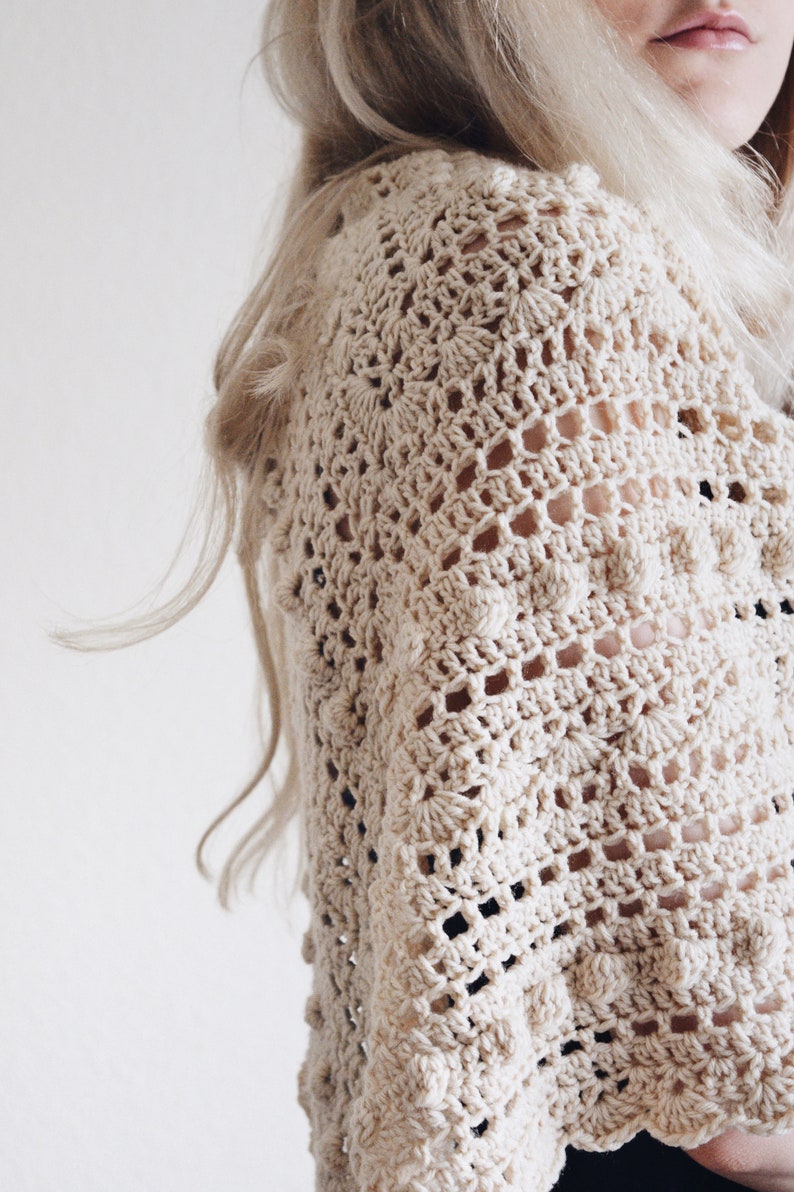 Woman wears a lace and bobble crochet scarf, The Isla Scarf, which was crocheted in cream yarn. This crochet shawl features lace and bobbles. [crochet triangle scarf, triangle shawl crochet patterns, beginner crochet, easy crochet patterns]