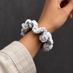 May include: A light gray scrunchie made of a soft, plush, velvet-like yarn with a textured, crocheted appearance. The accessory features a gathered, ruffled design that creates a voluminous, circular shape around the wrist.