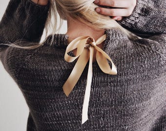 May include: A dark brown knitted sweater featuring a ribbed crew neckline and long sleeves. A smooth, light beige satin ribbon is tied into a bow at the center of the back neckline. The text DarlingJadore is visible in the bottom right corner.