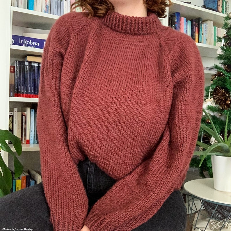 Woman wears a red knitted sweater, The Weekend Sweater, while sitting in front of a bookcase with plants. The pullover sweater is knitted in worsted weight yarn and this jumper looks cozy, featuring a double-knit neck and classic knit stitches.