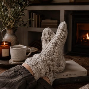 May include: A pair of thick, oatmeal-coloured knit socks featuring a vertical ribbed pattern and subtle brown flecks throughout the yarn. The socks are worn on the feet, extending up to the mid-calf, showcasing a textured, chunky knit construction.
