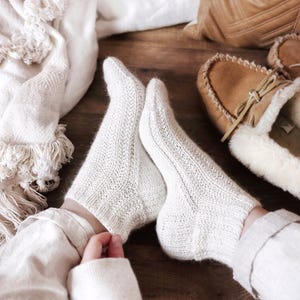 May include: Close-up of white knitted socks with detailed patterns. The socks are worn with a neutral-toned outfit, on a wooden surface. A blanket, pillow, and slippers are also visible, creating a warm, comfortable atmosphere.