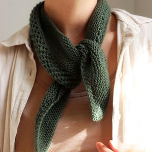 Puede incluir: Bufanda de punto verde oscuro atada al cuello. La bufanda está hecha de una tela de punto texturizada y está anudada en la parte delantera. La bufanda se usa sobre una camisa beige.