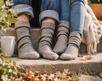 May include: Two pairs of feet wearing thick, light brown knit wool socks with a ribbed cuff design. The socks are mid-calf length and feature a textured, hand-knit appearance. The feet are positioned together on a concrete step, showcasing the soft, cozy material and the neutral earth-toned color of the yarn used in the construction of these warm winter garments.