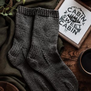 May include: A pair of dark charcoal gray knit socks featuring a ribbed cuff and a textured heel detail. The socks are displayed alongside a small framed sign with the text "CABIN SWEET CABIN" and a graphic of a mountain cabin and pine trees.