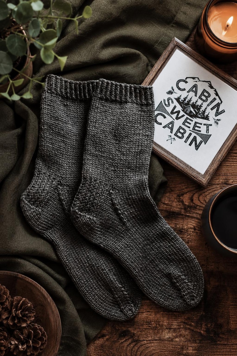 May include: A pair of dark charcoal gray knit socks featuring a ribbed cuff and a textured heel detail. The socks are displayed alongside a small framed sign with the text "CABIN SWEET CABIN" and a graphic of a mountain cabin and pine trees.
