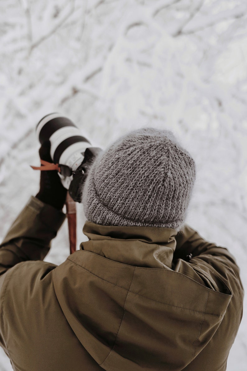 May include: A grey knit beanie worn with an olive green hooded jacket. The individual is holding a professional camera with a white lens, wearing dark brown gloves, and has a thin red strap attached to the camera body for secure handling.