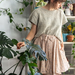 Woman wears a knitted summer tshirt, The Classic Tee, in a room of plants. The knit shirt features a raglan construction and simple knit stitches (Stockinette). This basic knit tee tshirt looks lightweight and comfortable to wear.
[knitting patterns]