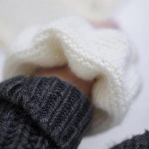 May include: A dark charcoal gray knitted sweater featuring a ribbed cuff at the wrist. A soft, white, chunky knit fabric is layered over the sleeve, showcasing a distinct contrast in texture and color between the two knitted garments.