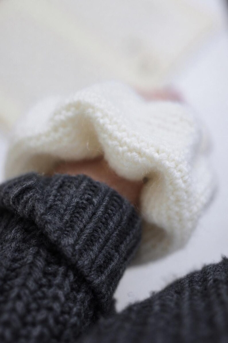 May include: A dark charcoal gray knitted sweater featuring a ribbed cuff at the wrist. A soft, white, chunky knit fabric is layered over the sleeve, showcasing a distinct contrast in texture and color between the two knitted garments.