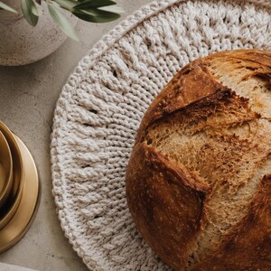 May include: A round, off-white crocheted placemat featuring a thick, textured cord pattern. A large, golden-brown crusty loaf of bread sits on the right side of the placemat, showcasing a deep, scored cross pattern on its rounded top surface.