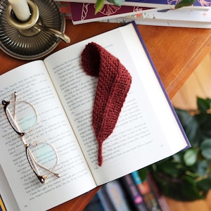 May include: A dark red knitted bookmark shaped like a leaf resting on an open book. The bookmark features a textured garter stitch pattern and a pointed tip. Beside the book, a pair of thin-rimmed glasses sits on the page, and a candle is nearby.