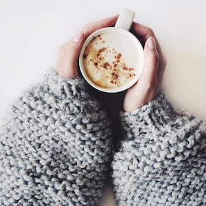 May include: A white ceramic mug filled with a creamy coffee beverage topped with a light dusting of brown cinnamon powder. The mug is held by two arms wearing a thick, chunky-knit sweater in a heathered light gray wool yarn with visible texture.